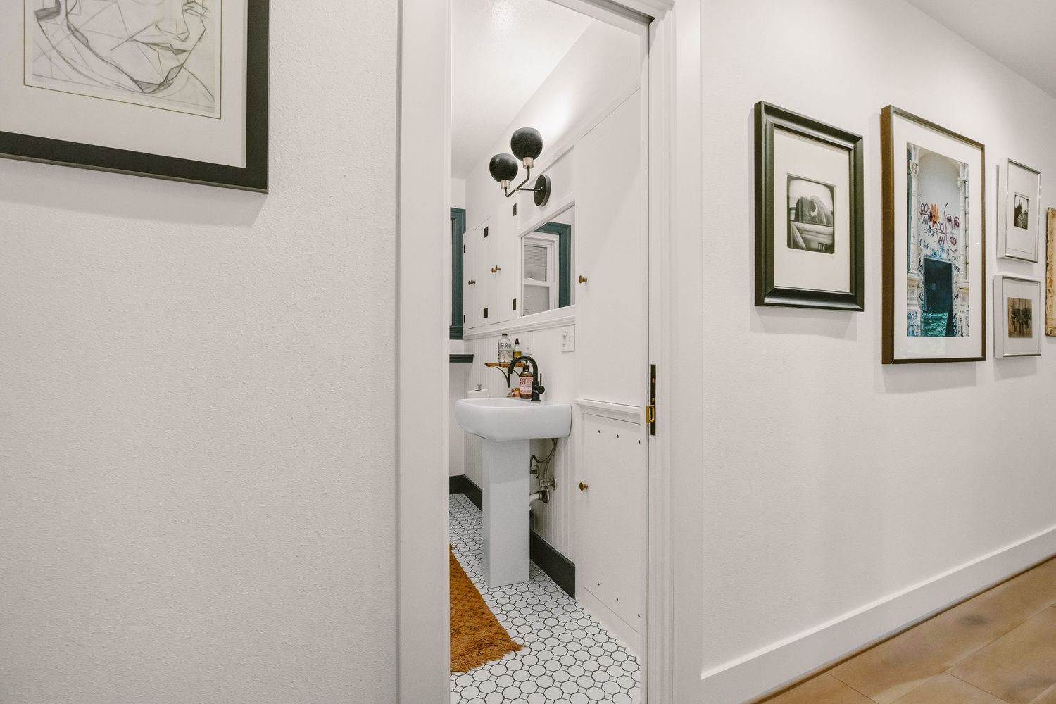 Elegant hallway leading to a white pedestal sink bathroom with black accents, mosaic tile floor, and framed wall art.