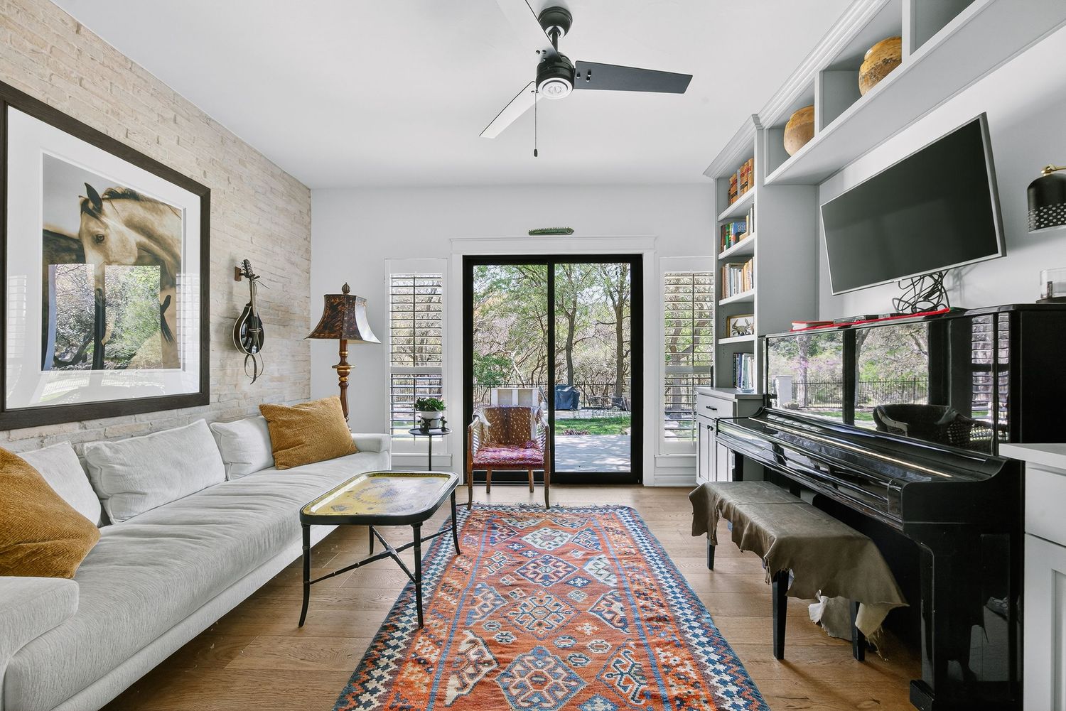 Bright living room with white sofa, colorful rug, piano, built-in shelves, ceiling fan, and French doors to patio.