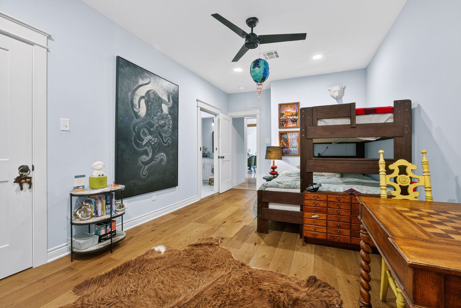 Kids bedroom with hardwood floors, bunk bed, ceiling fan, large wall art, and a cozy brown rug in a bright space.