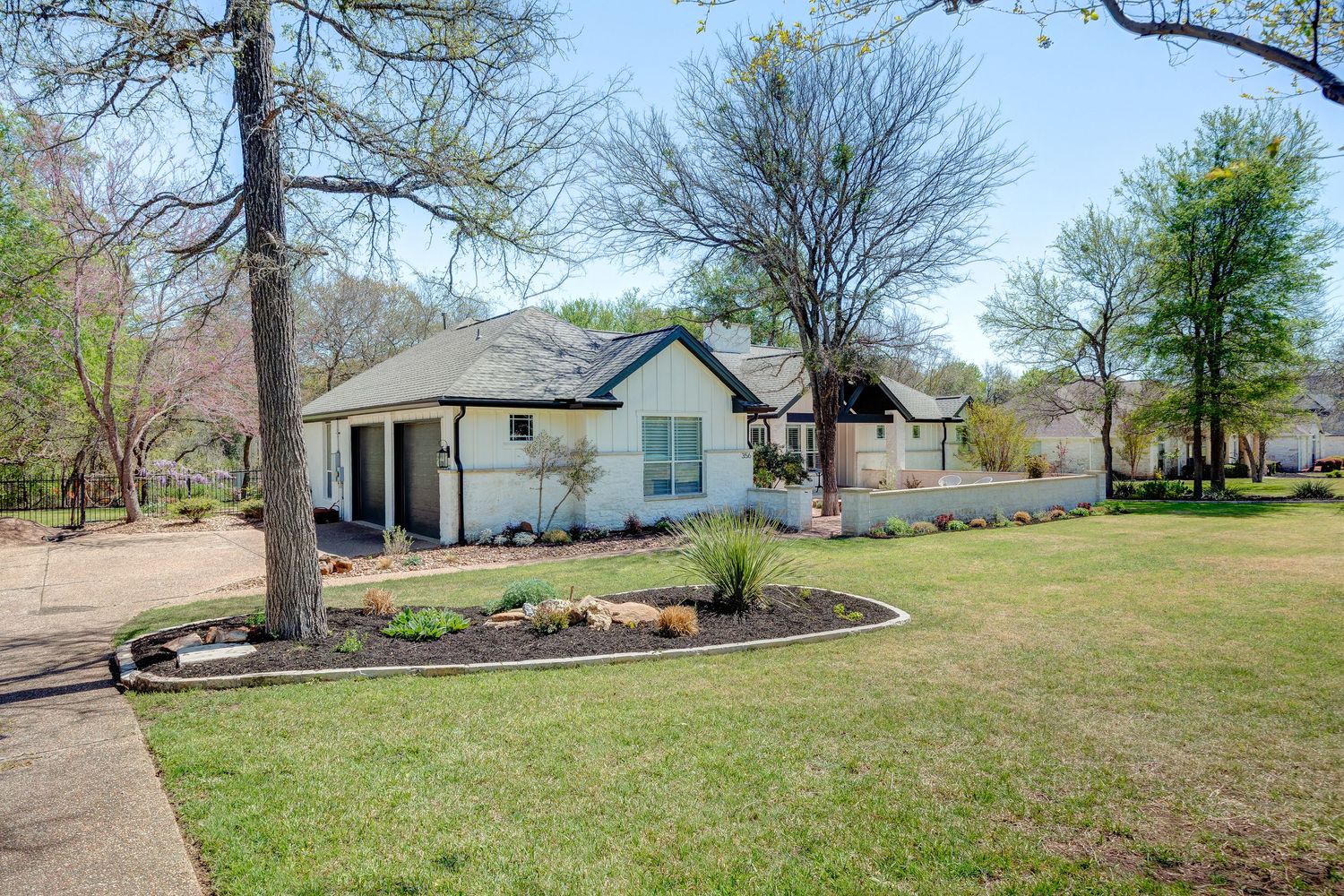 White ranch-style home with manicured lawn, mature trees, and landscaped garden beds on a sunny spring day.
