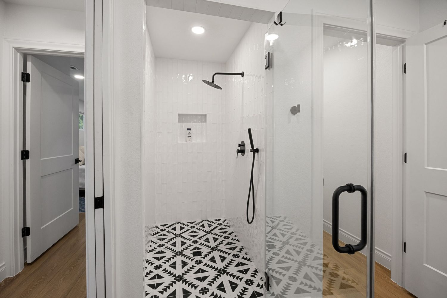 Modern walk-in shower with geometric black and white patterned tile floor, white subway walls, and matte black fixtures behind glass door.