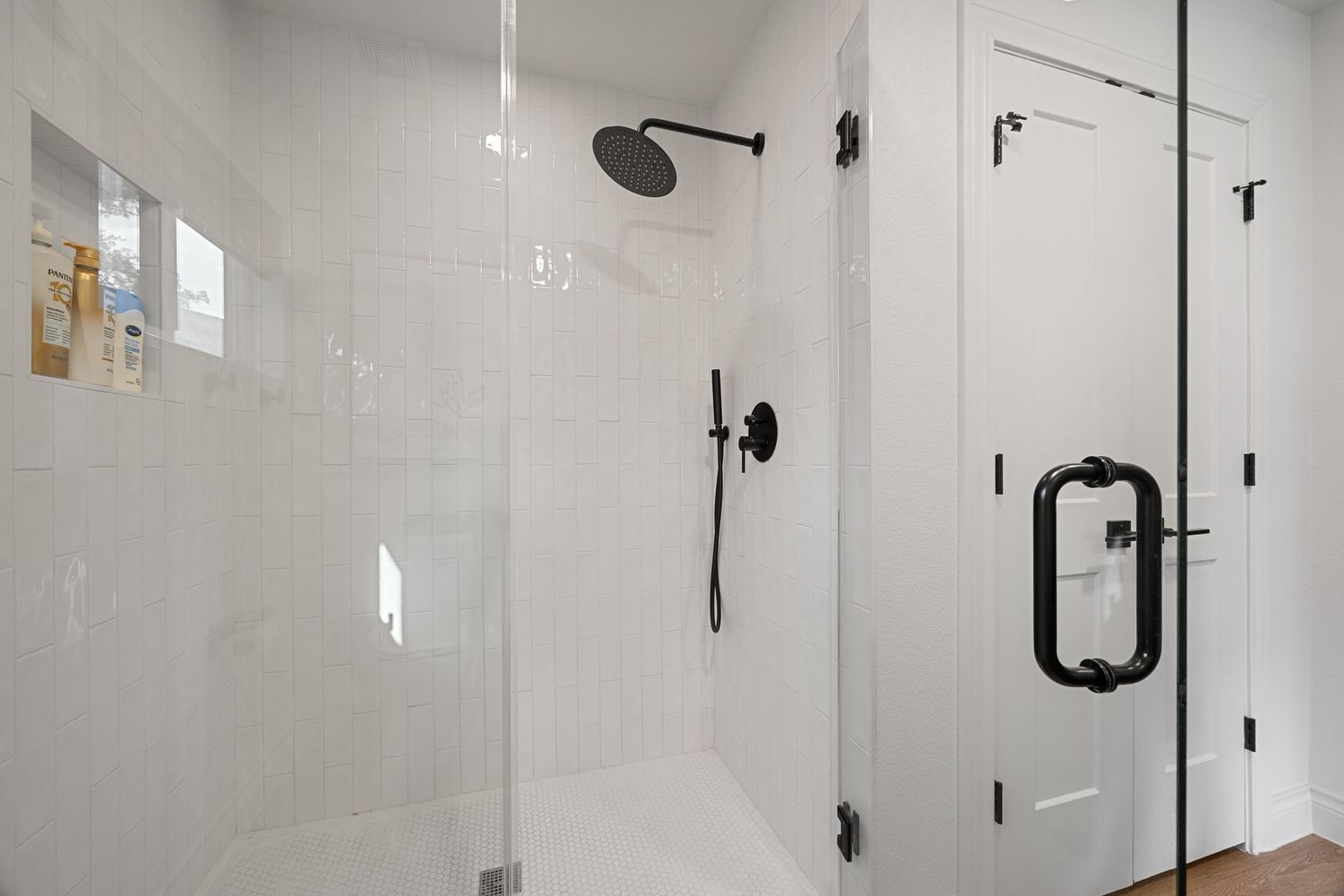 Modern walk-in shower with white subway tiles, black rainfall showerhead, handheld fixture, and frameless glass door.