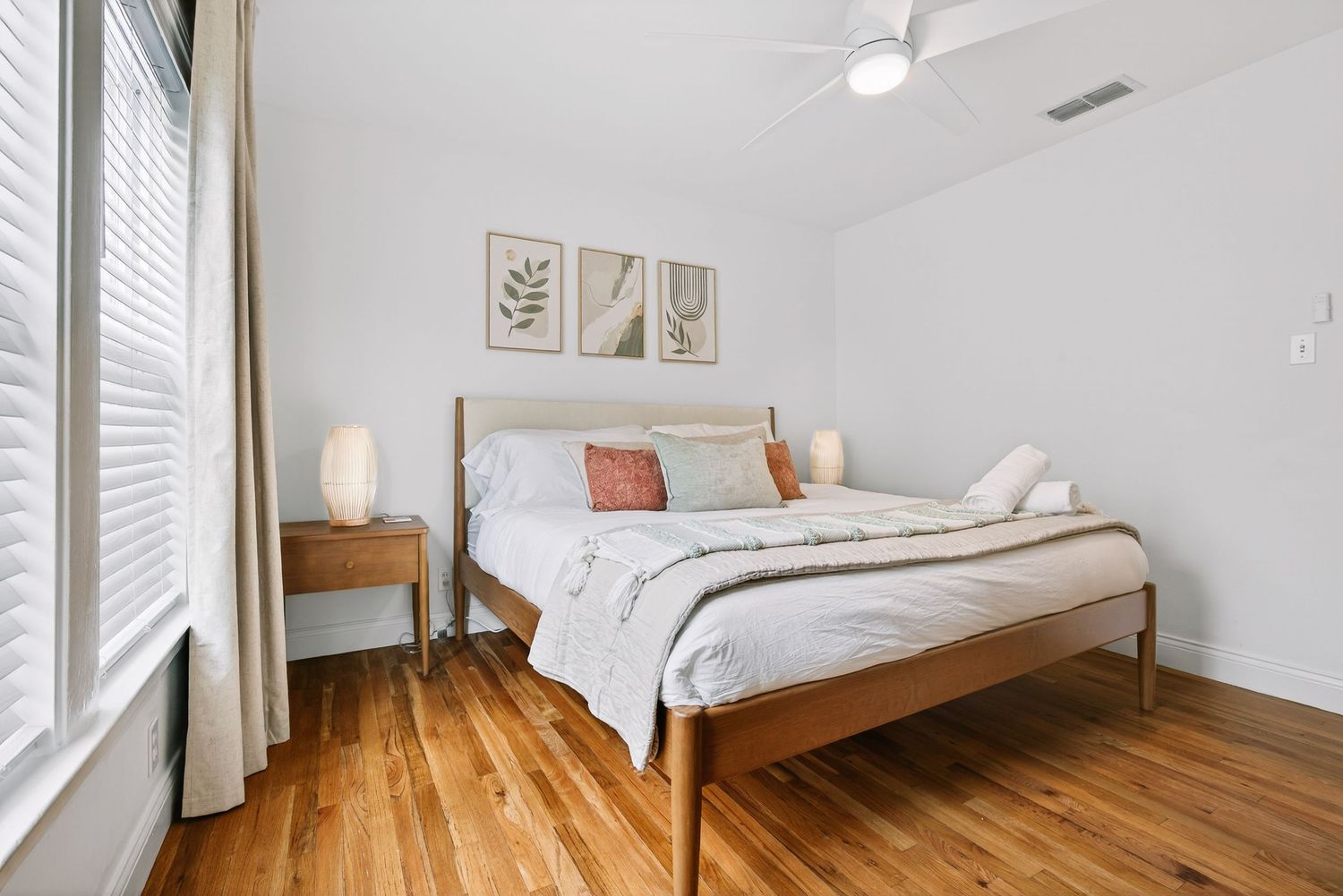 Modern bedroom with wooden floors, white walls, minimalist decor, and natural light streaming through window blinds.