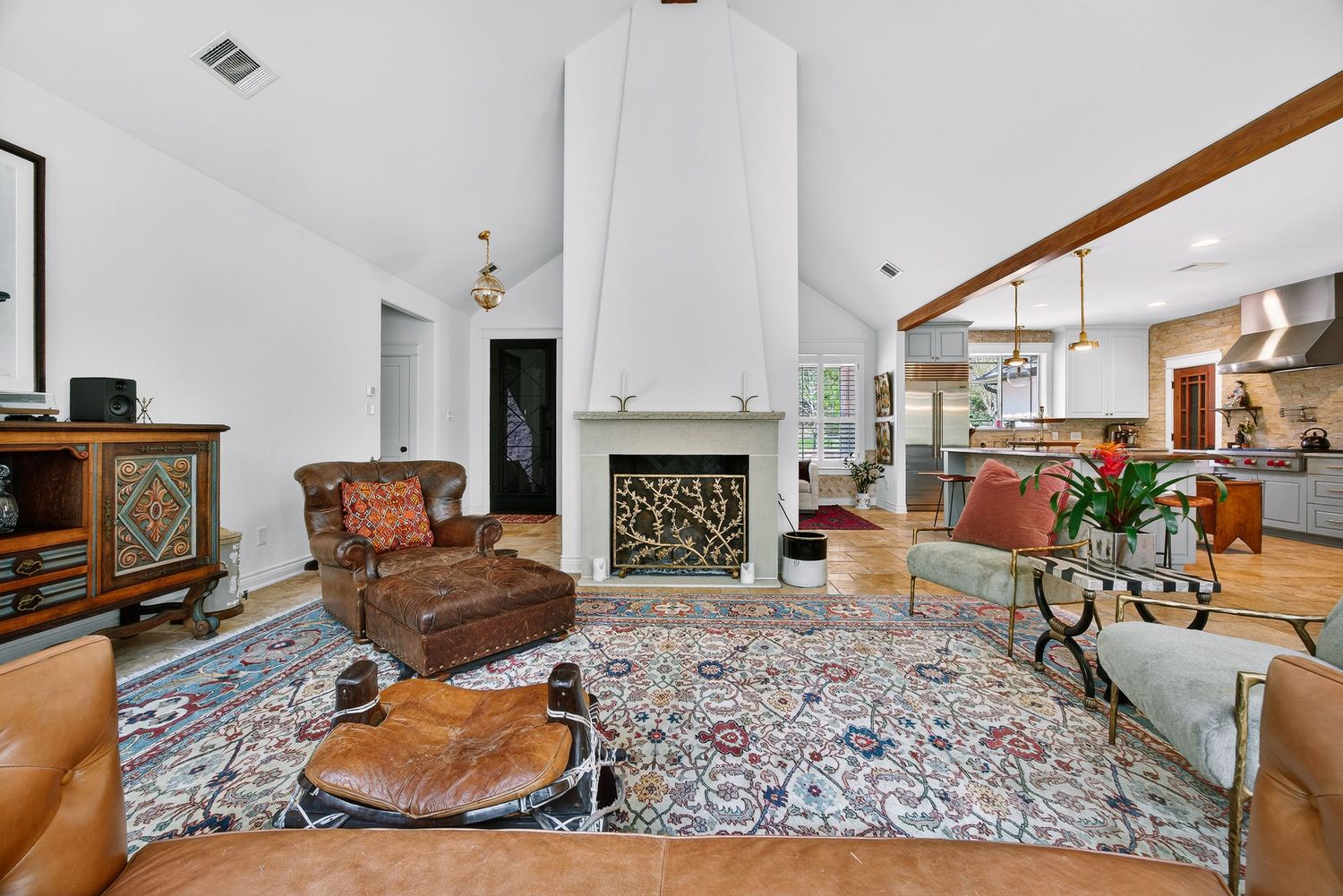 Spacious living room with vaulted ceilings, white fireplace, ornate area rug, leather furniture, and open kitchen in background.
