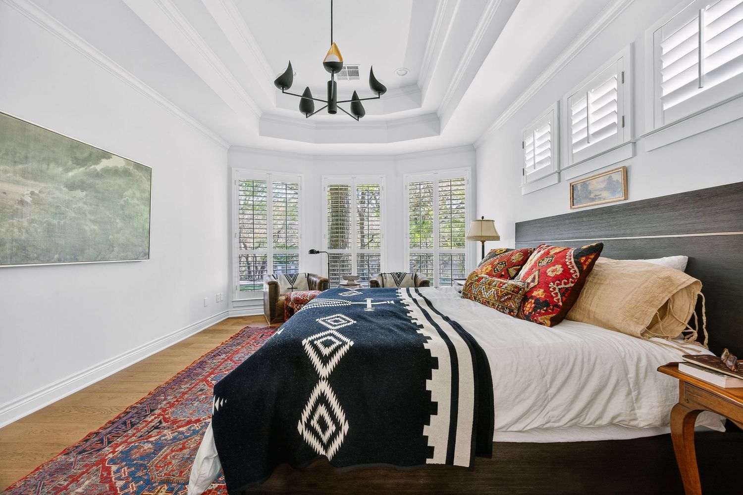 Elegant bedroom with tray ceiling, chandelier, hardwood floors, patterned bedding, and shuttered bay windows.