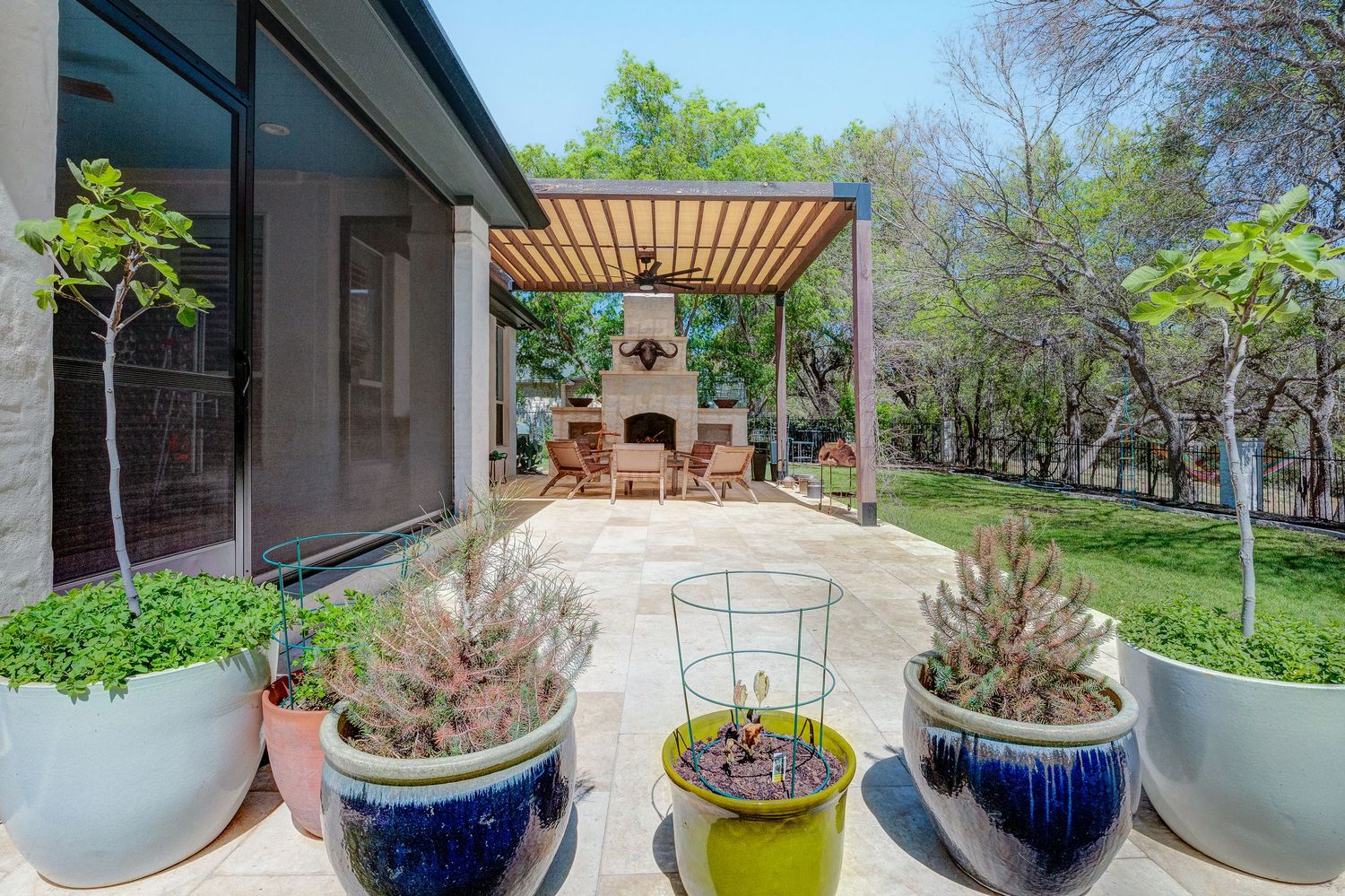 Outdoor patio with pergola, stone fireplace, seating area, and colorful potted plants surrounded by lush green trees.