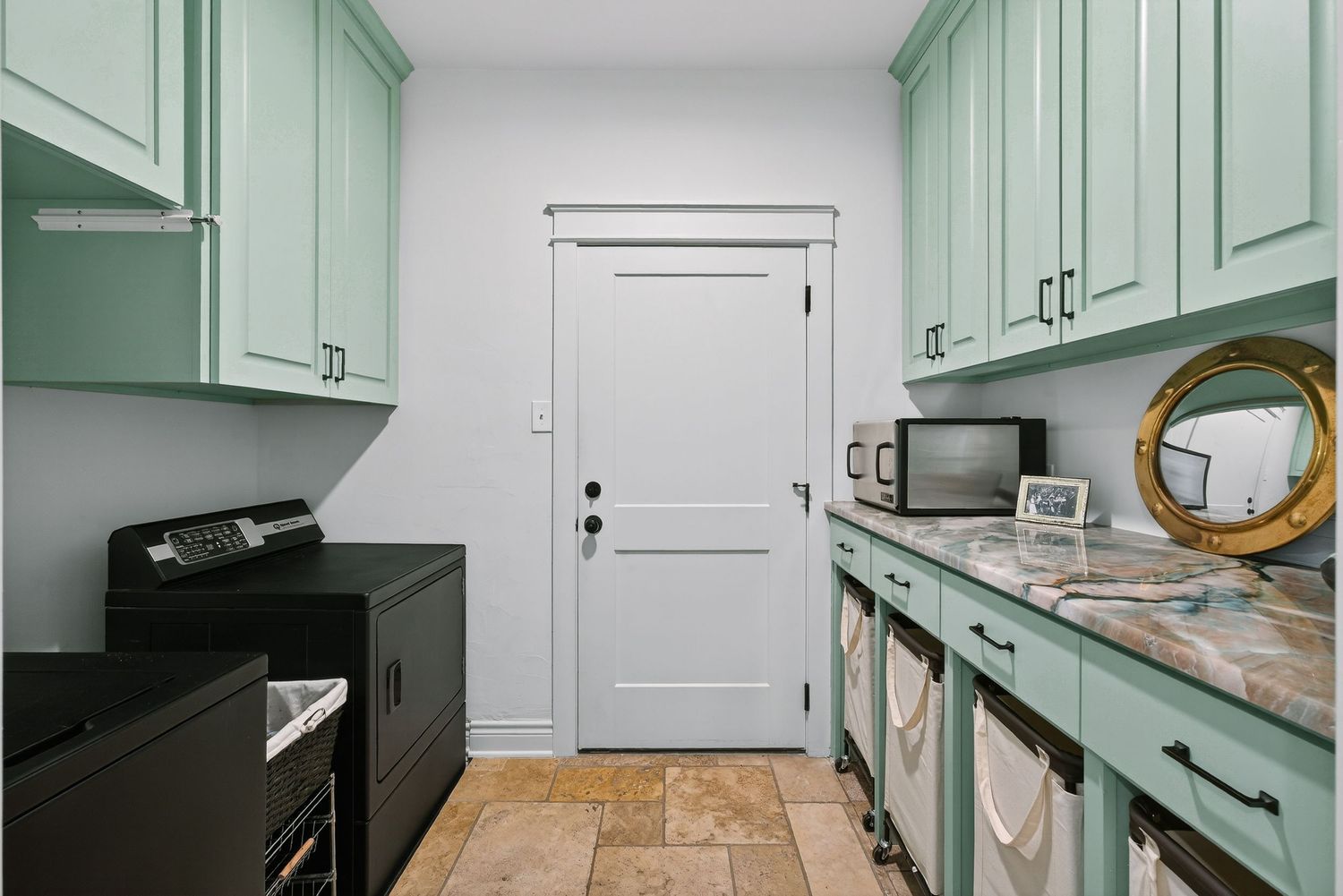 Laundry room with mint green cabinets, marble countertop, black washer and dryer, and white door on tile floors.