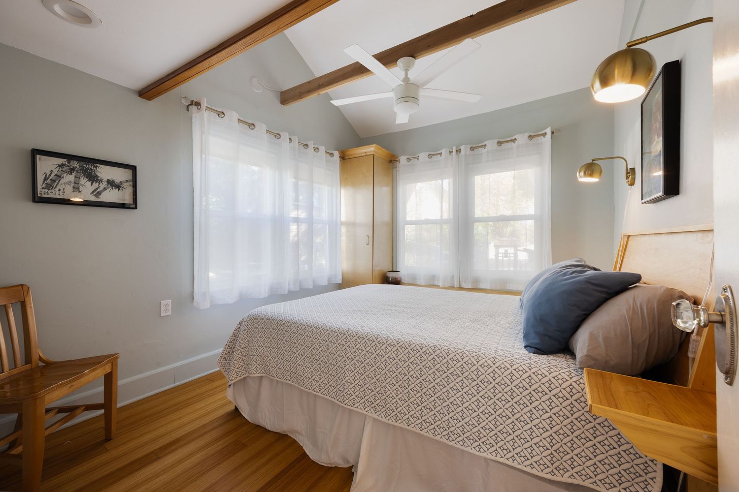 Cozy bedroom with white bedding, wooden beams, gold sconces, and sheer curtains in a modern coastal style.