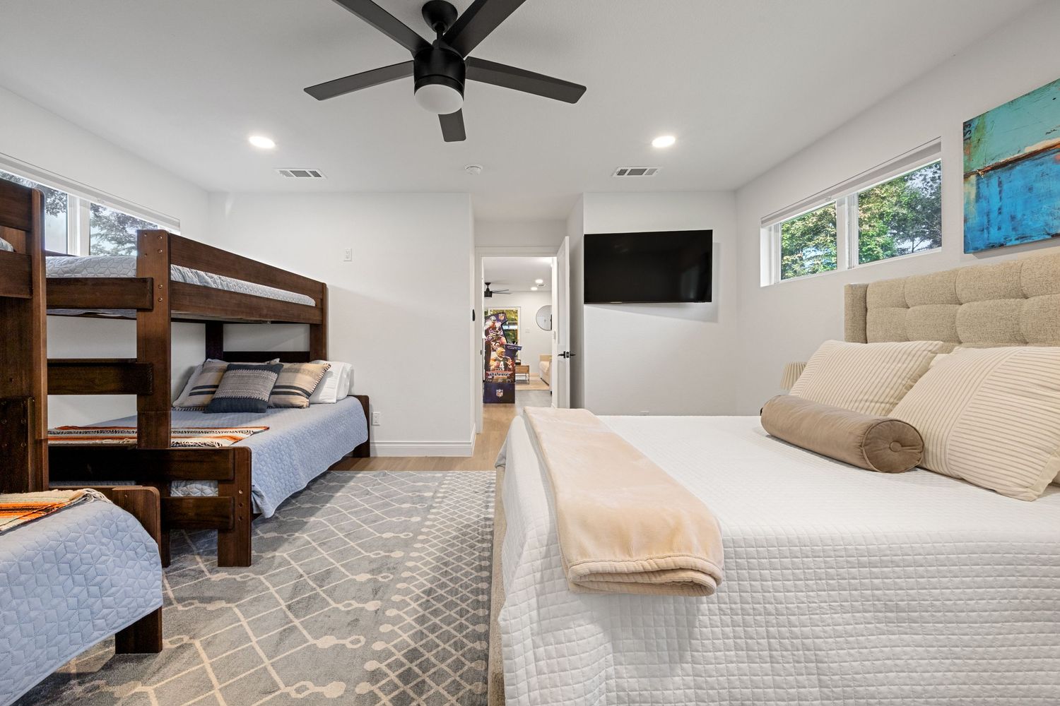 Bright bedroom with bunk beds, queen bed, ceiling fan, wall-mounted TV, and multiple windows for natural light.