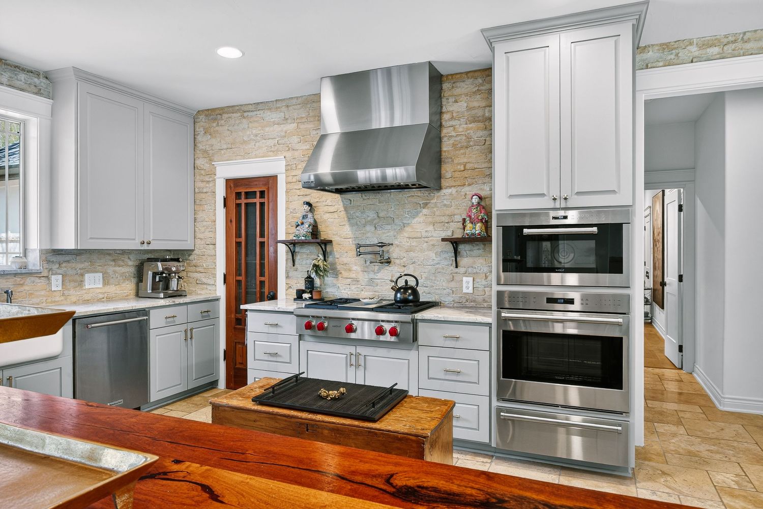 Modern kitchen with white cabinets, stainless steel appliances, stone backsplash, range hood, and rustic wood countertop accents.