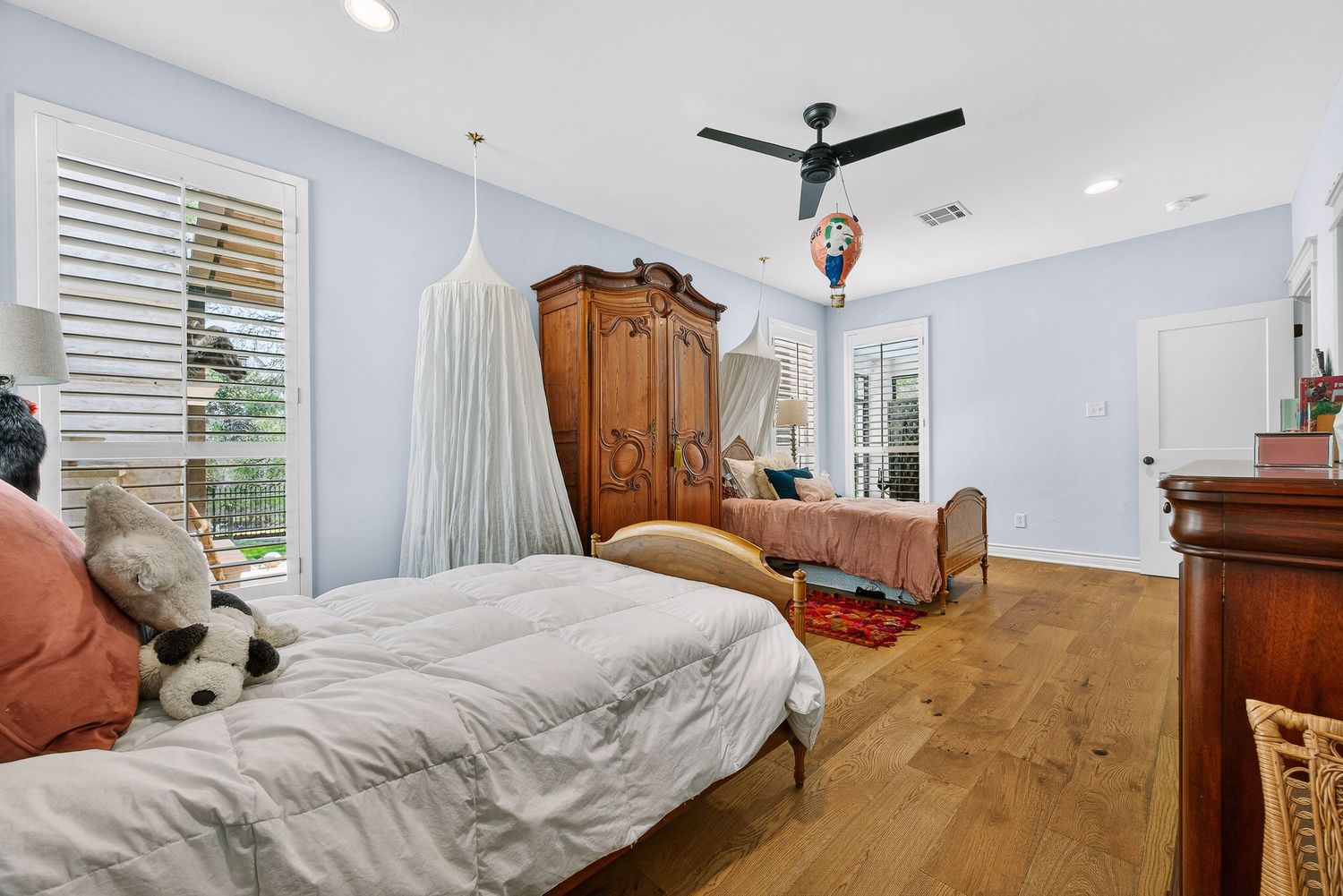 Bright bedroom with hardwood floors, ceiling fan, antique armoire, canopy bed, and plantation shutters on windows.