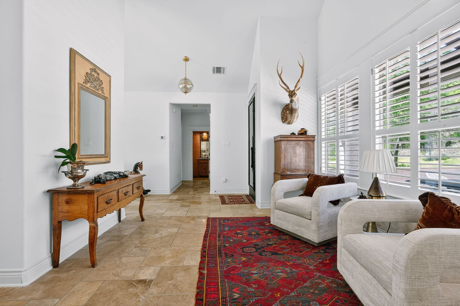 Elegant living room with travertine floors, red Persian rug, white sofa, antler mount, plantation shutters, and a wooden console table.