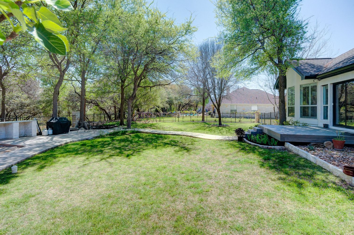 Spacious backyard with lush green lawn, mature trees, stone patio, and a deck adjacent to a home on a sunny day.