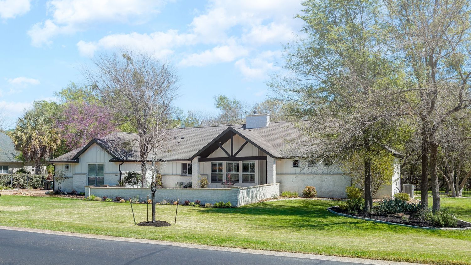 Spacious single-story ranch home with white exterior, wood accents, manicured lawn, and mature trees on a sunny day.