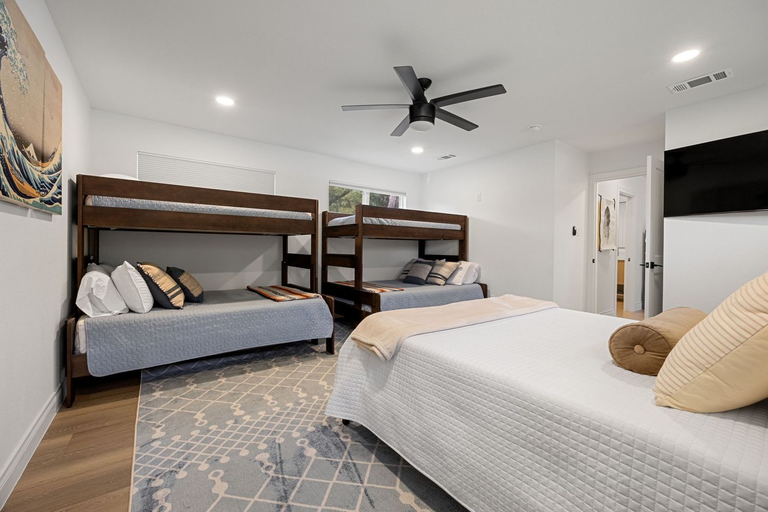 Spacious bedroom with two bunk beds, a queen bed, ceiling fan, wall-mounted TV, and patterned area rug on hardwood floors.