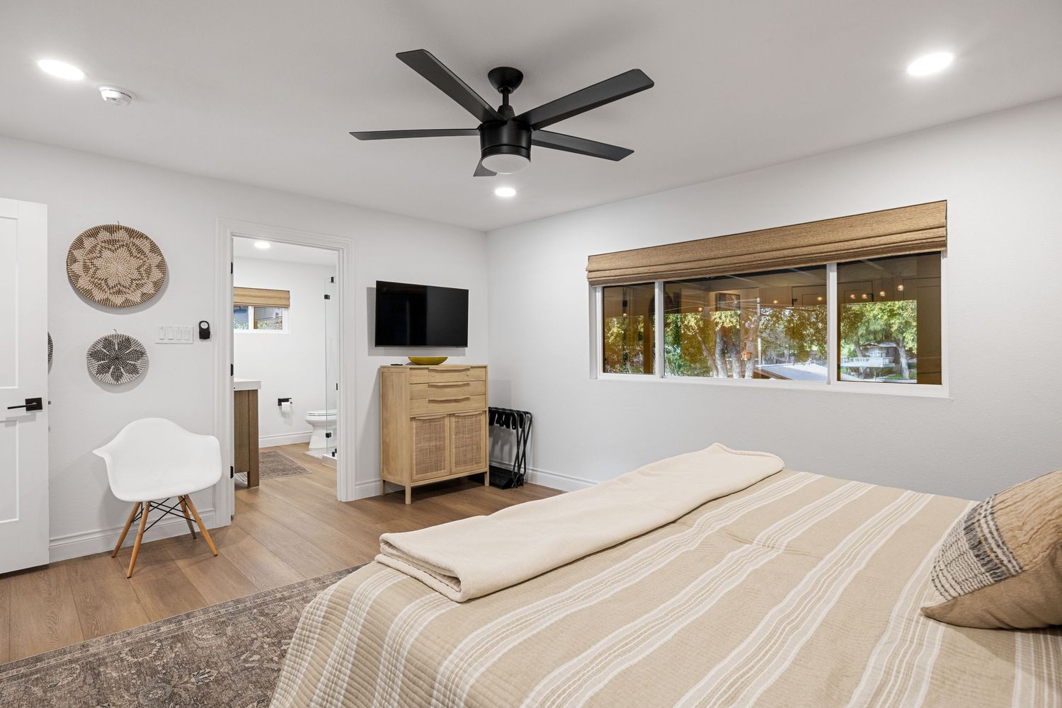 Bright bedroom with ceiling fan, wall-mounted TV, wood dresser, and natural light from large window.