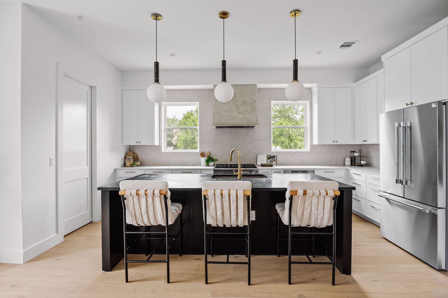 Modern white kitchen with black island, gold pendant lights, bar stools, stainless fridge, and hardwood floors.