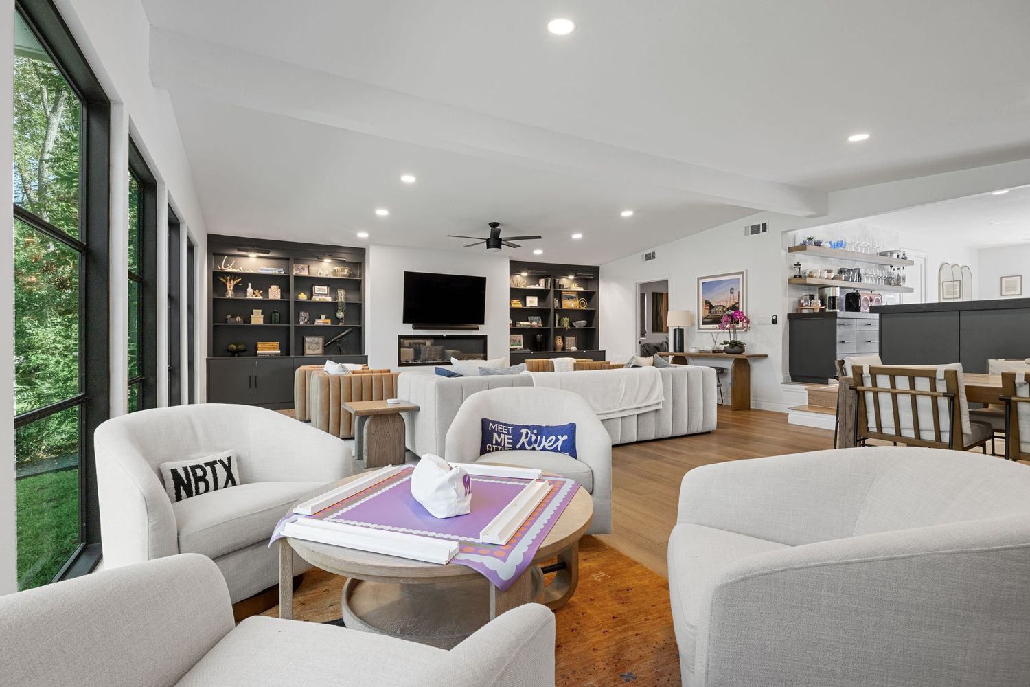 Spacious open-concept living room with white sofas, built-in shelves, wall-mounted TV, hardwood floors, and large windows.