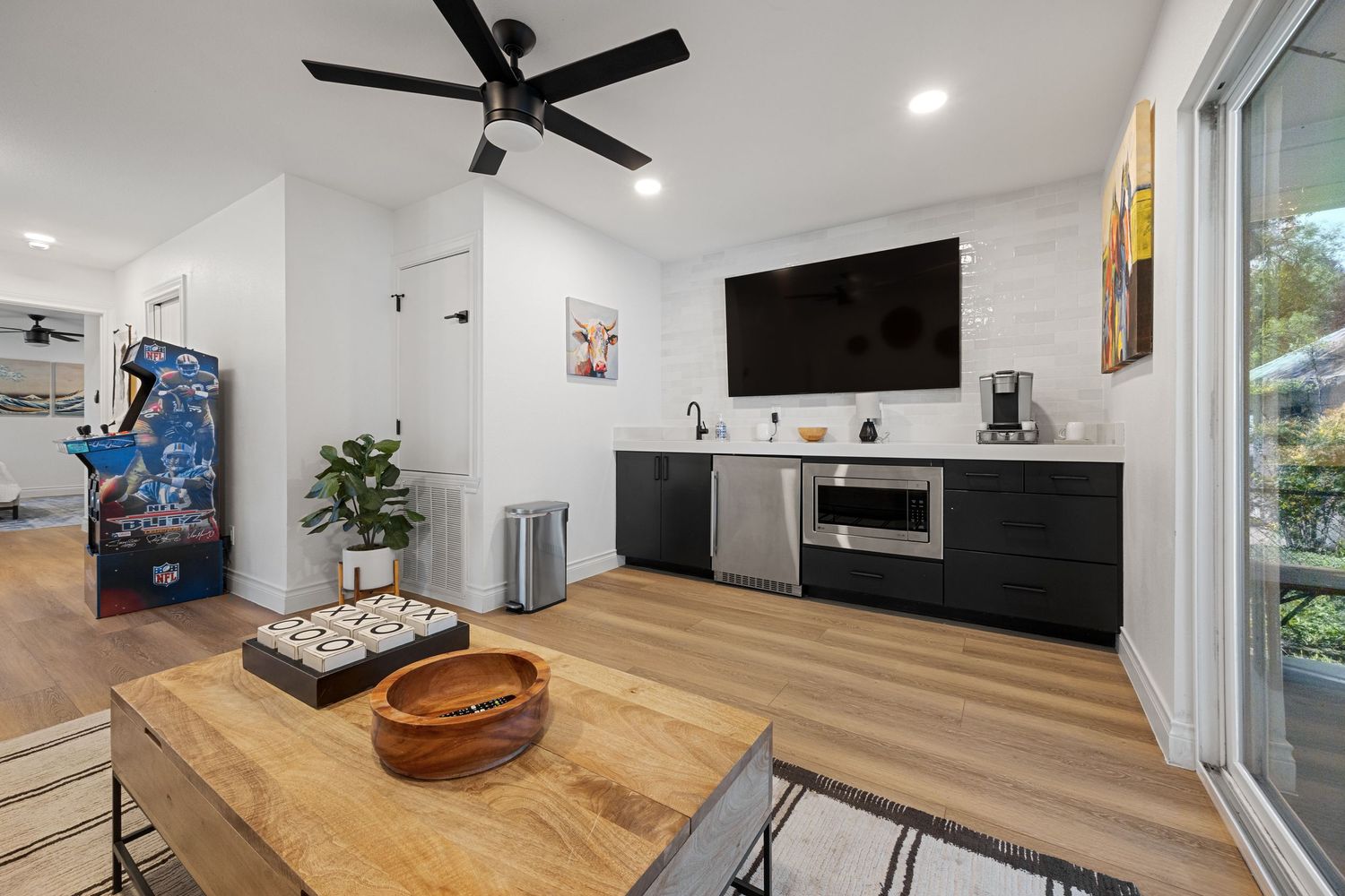 Contemporary kitchenette with dark cabinetry, stainless steel appliances, wall-mounted TV, light hardwood floors, and wooden dining table.