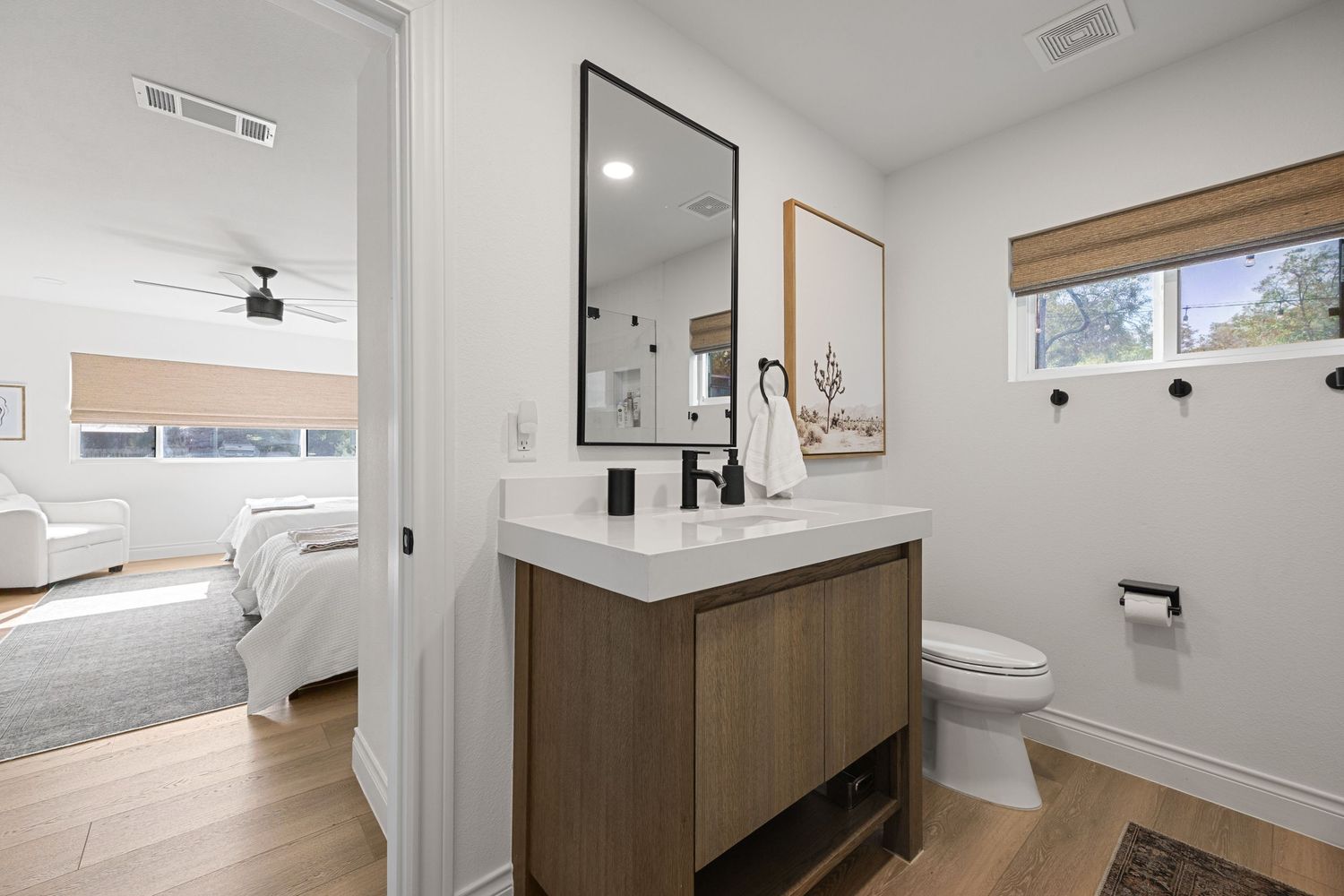 Modern bathroom with wood vanity, white countertop, black-framed mirror, and view into bright bedroom.