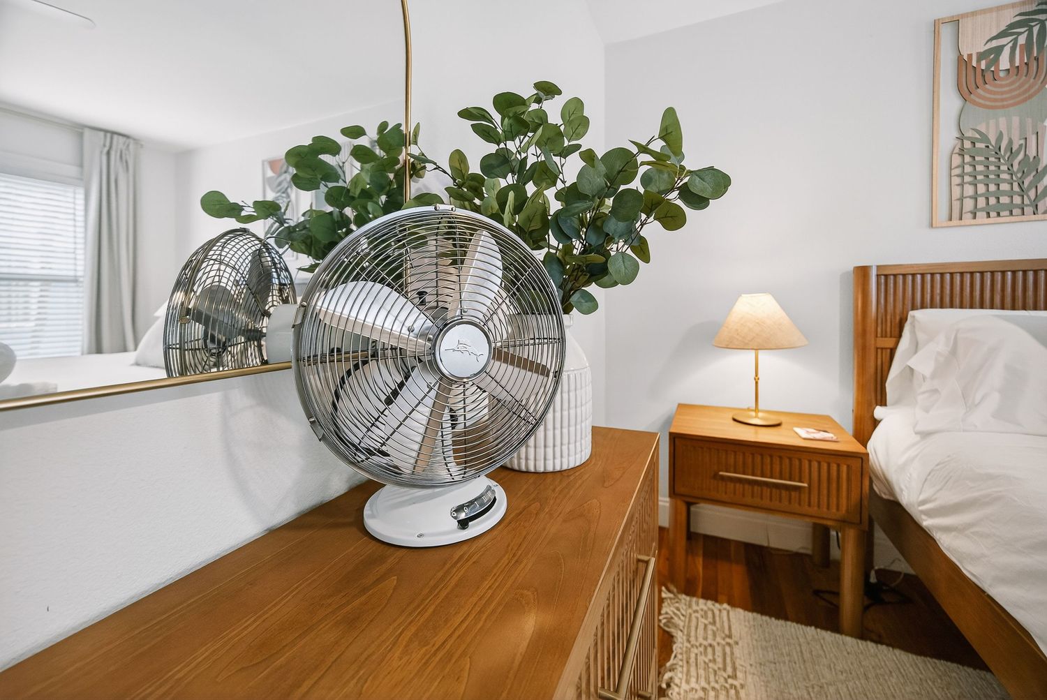 White bedroom with hardwood floors, wooden furniture, houseplants and a standing fan.