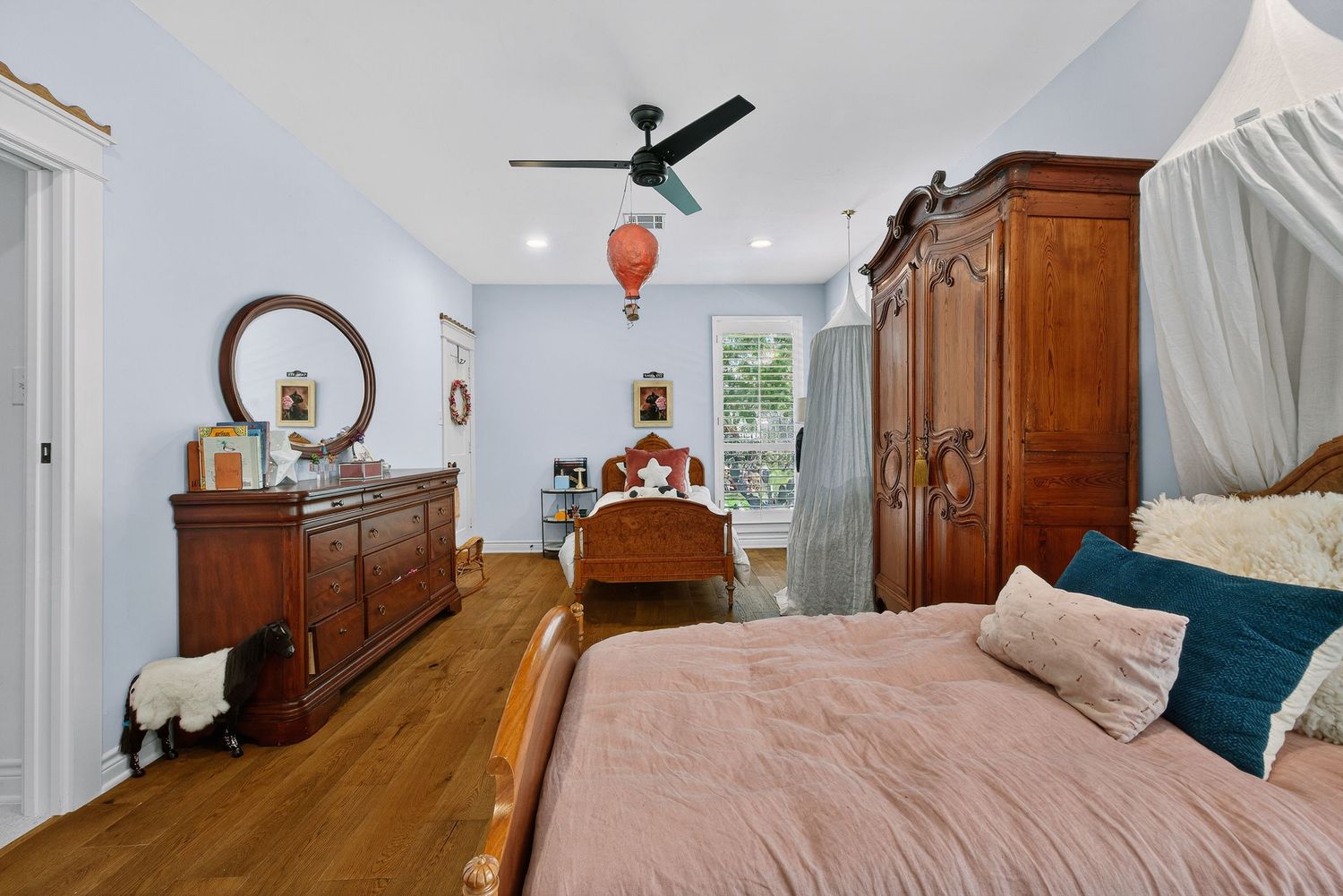 Spacious bedroom with hardwood floors, ornate wooden armoire, ceiling fan, and a cozy bed with pink bedding.
