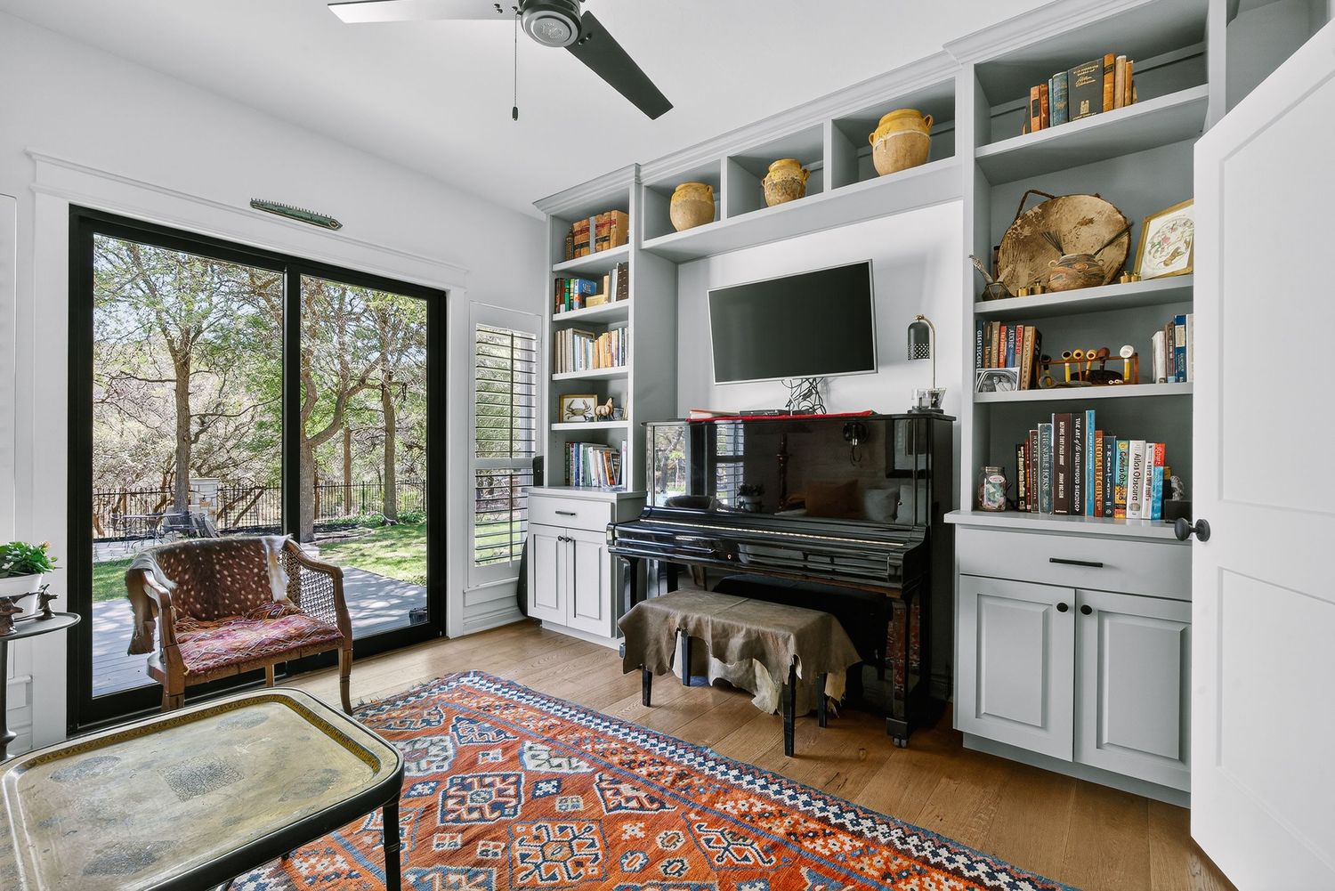 Cozy home office with built-in gray shelves, piano, wall-mounted TV, Persian rug, and sliding glass doors to a garden.