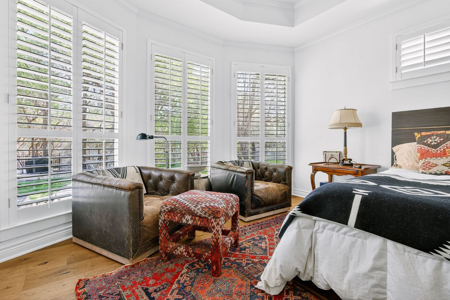 Elegant bedroom with plantation shutters, hardwood floors, leather chairs, and a vibrant Persian rug in a bright, airy space.