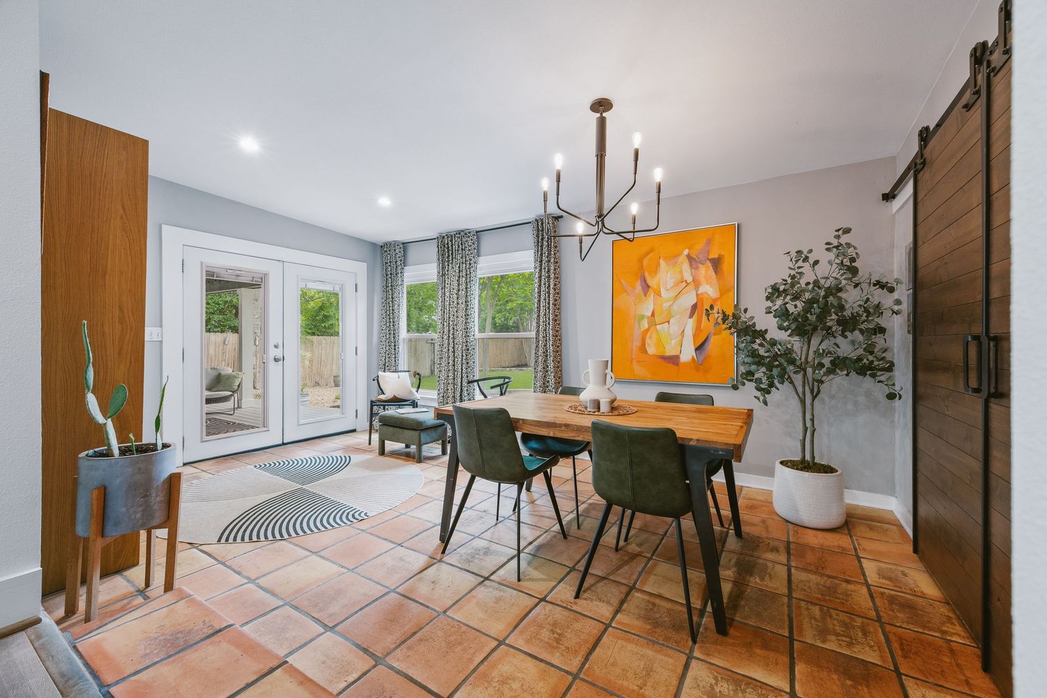 Bright dining room with terracotta tile floors, wooden table, green chairs, barn door, and French doors leading to a patio.