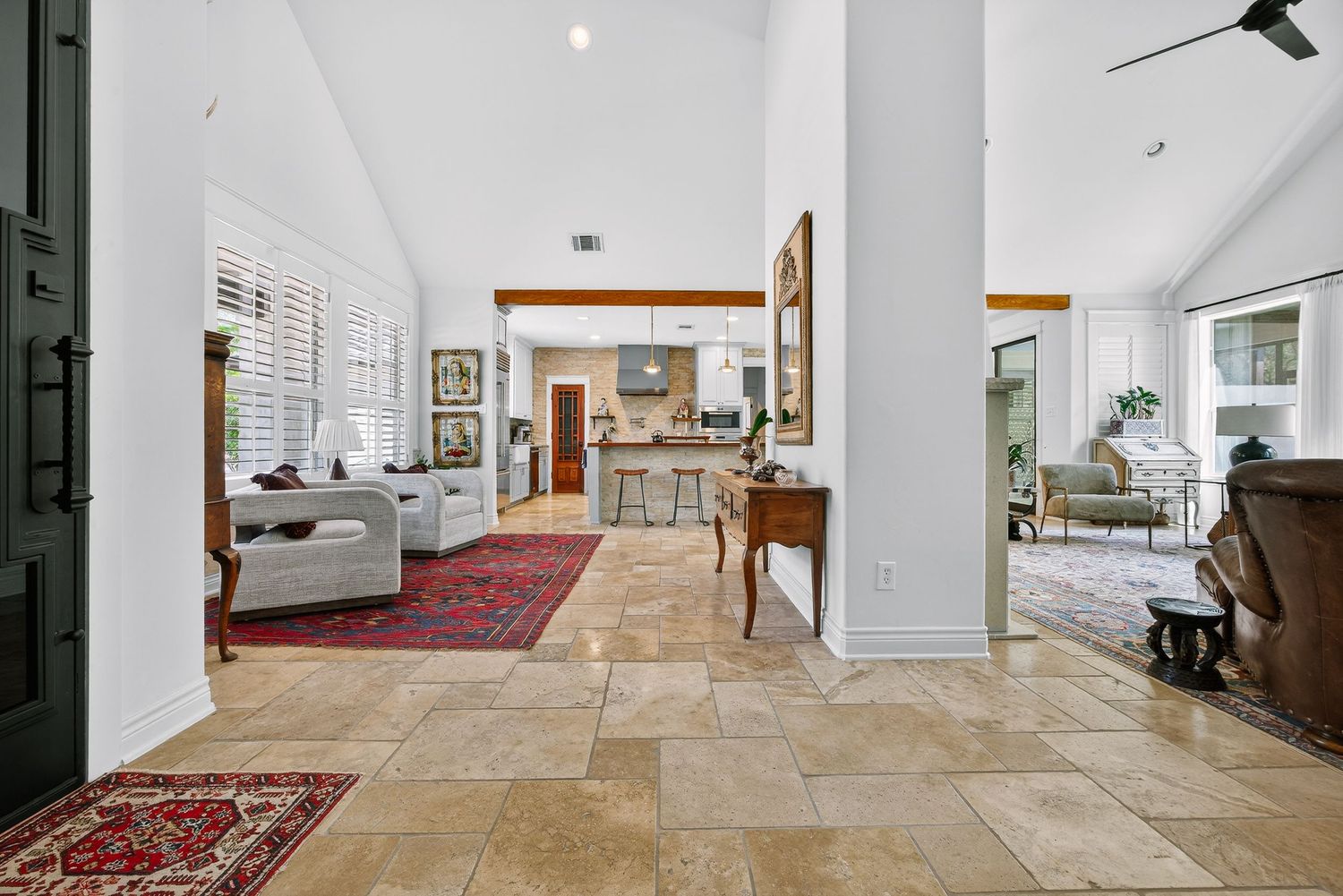 Spacious open-concept living area with travertine tile floors, high vaulted ceilings, red oriental rug, and view into modern kitchen.