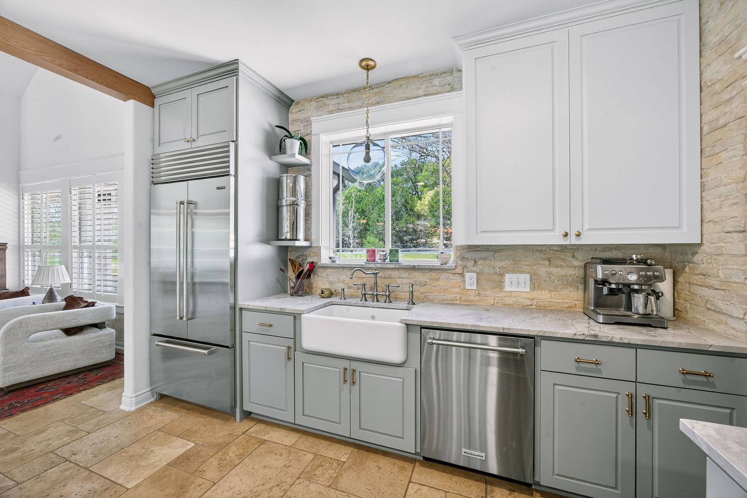 Bright kitchen with gray cabinets, farmhouse sink, stainless steel appliances, stone backsplash, and pendant lighting over tile floors.