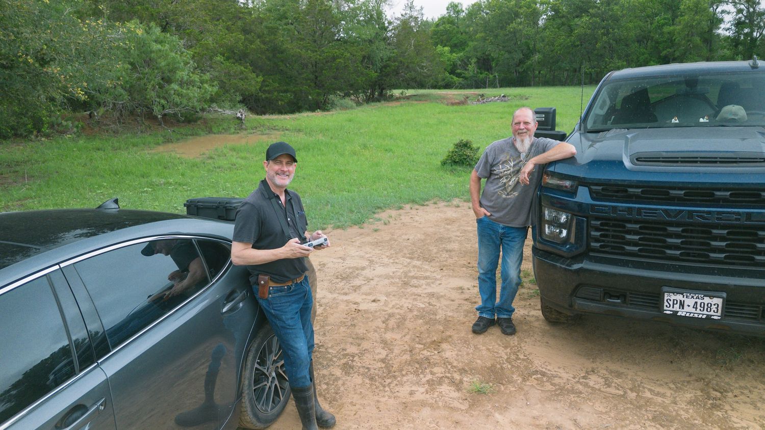 Two men standing between a black sedan and a dark pickup truck on a rural dirt path surrounded by green trees.