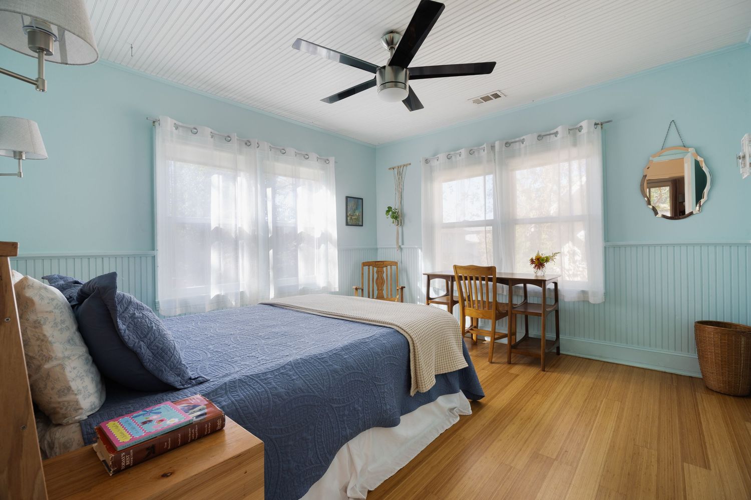 Light blue bedroom with hardwood floors, ceiling fan, and dining area by windows.