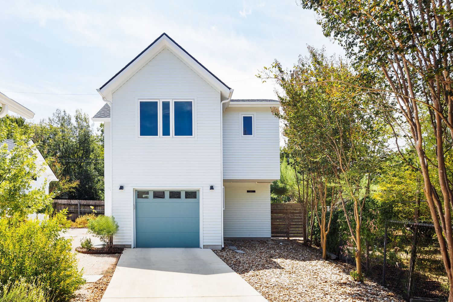 Modern two-story white home with teal garage door, blue-framed windows, concrete driveway, and mature trees in a landscaped yard.
