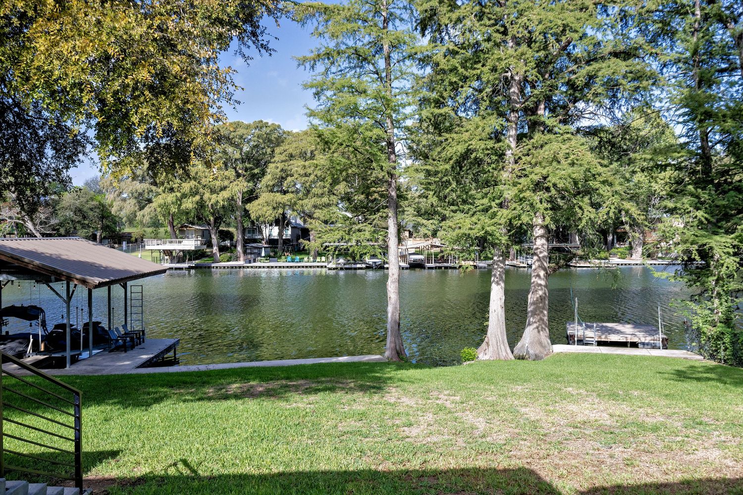 Serene lakeside view with lush green lawn, towering cypress trees, and private boat docks reflecting on calm water.