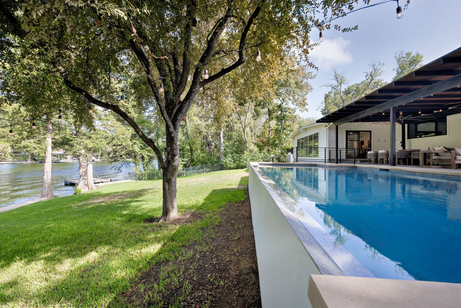 Modern infinity pool beside a glass house with lush green lawn and mature trees overlooking a serene waterfront.