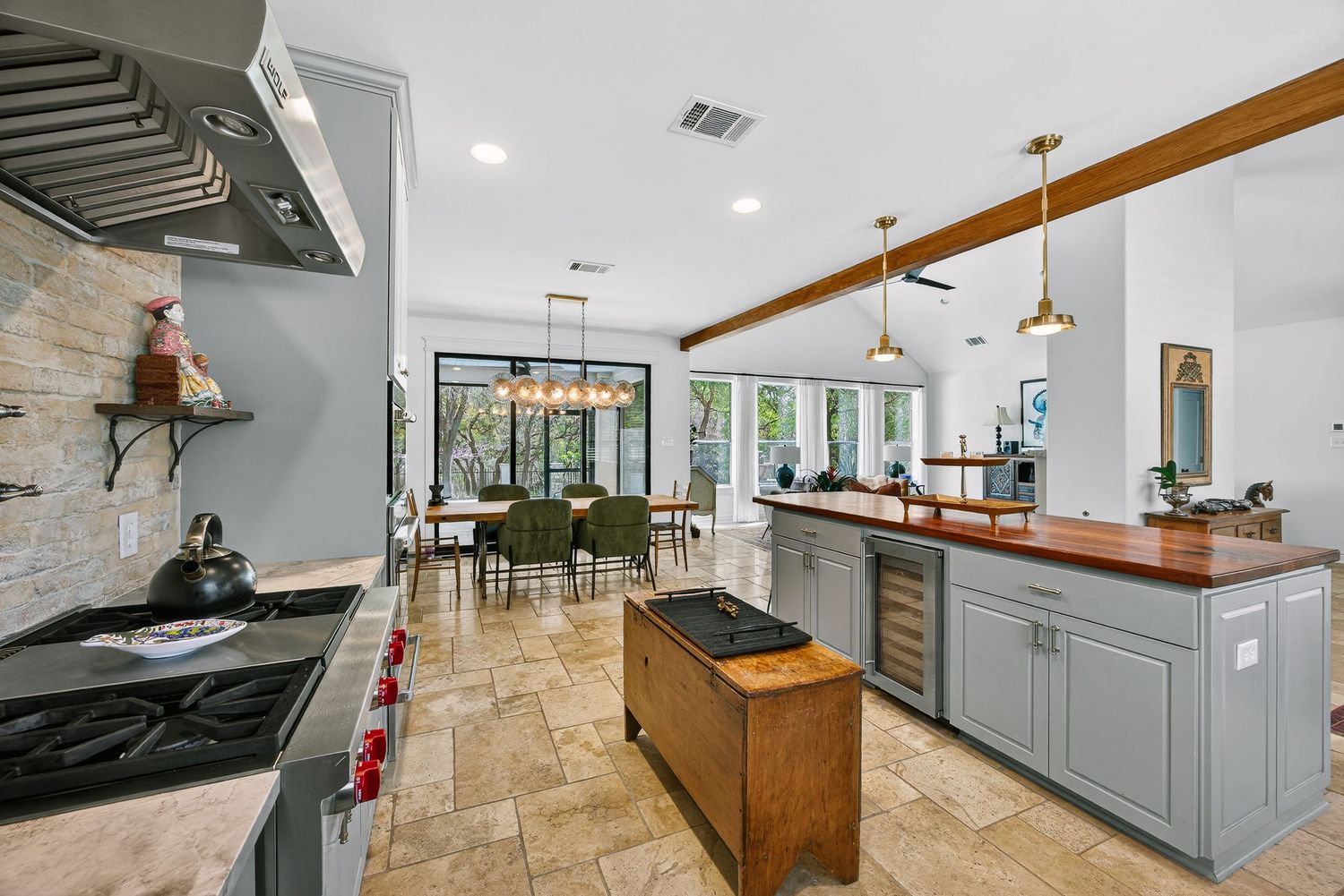 Modern kitchen with gray cabinets, wood beam ceiling, wine fridge, and stone tile floors opening to a bright dining area.
