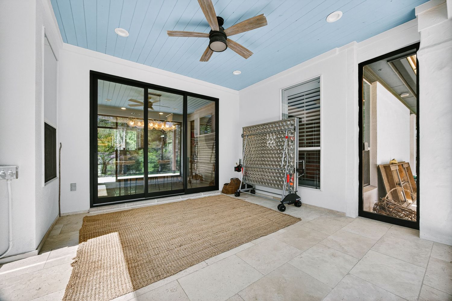 Bright enclosed patio with blue ceiling, ceiling fan, sliding glass doors, and tile flooring with a woven rug.