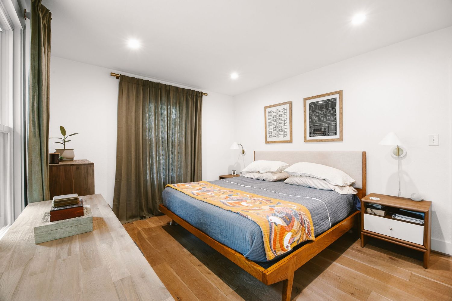 Modern mid-century bedroom with wooden bed frame, blue bedding, hardwood floors, and neutral white walls with recessed lighting.