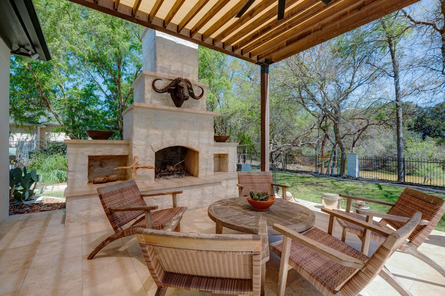 Covered outdoor patio with stone fireplace, wicker chairs, and pergola overlooking a lush green backyard.