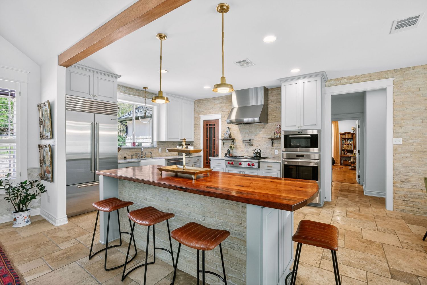 Modern kitchen with wood-topped island, gold pendant lights, gray cabinets, and bar stools on travertine tile floors.