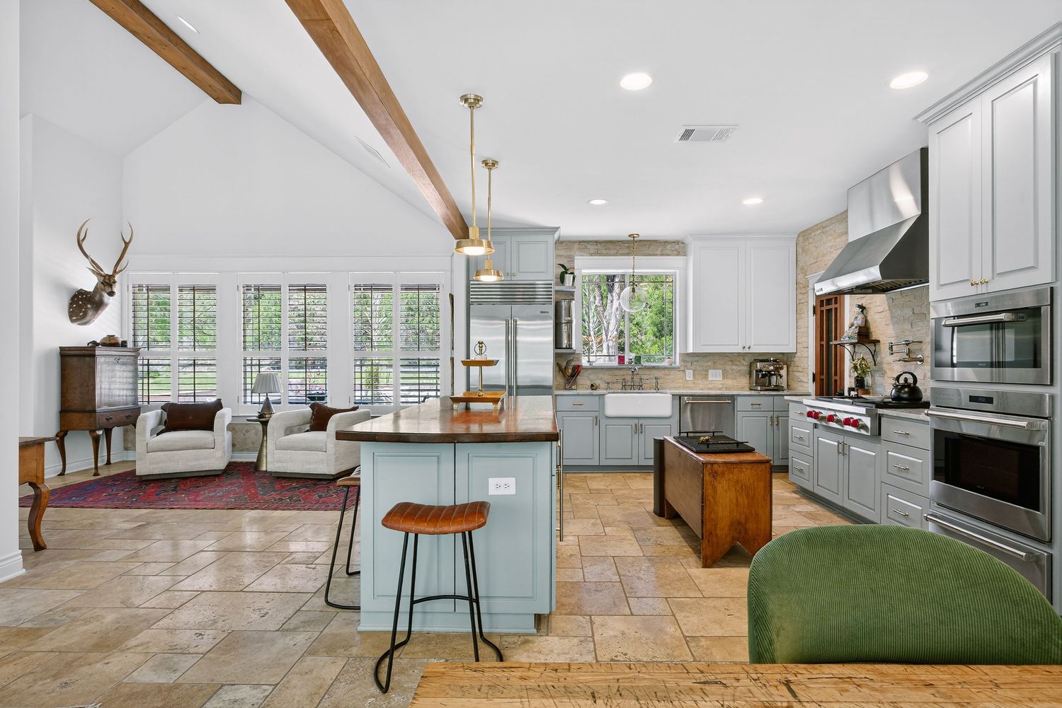 Open-concept kitchen with blue island, wood beams, pendant lights, gray cabinets, and a cozy living area with large windows.