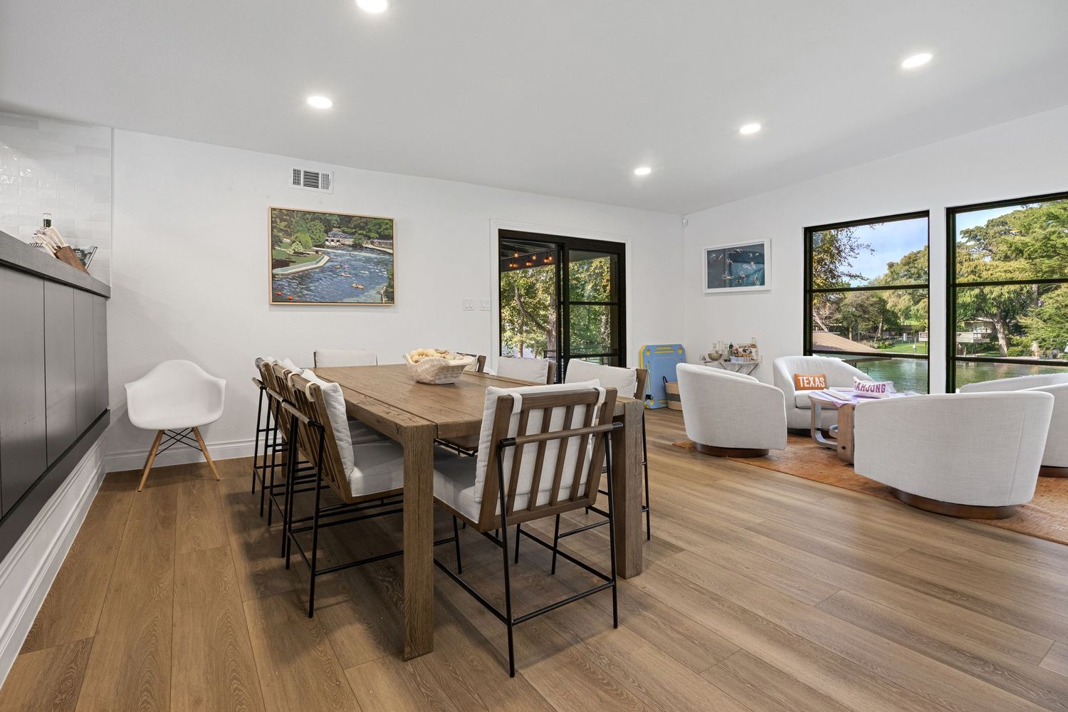 Modern dining room with wood floors, rectangular table, white chairs, and large windows overlooking greenery.