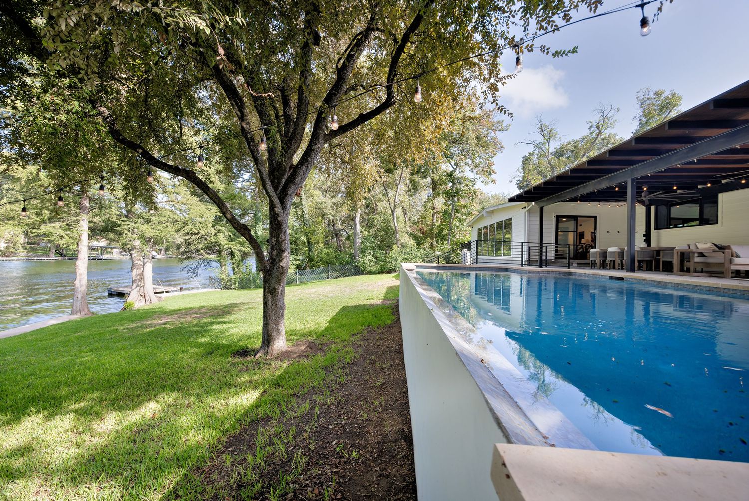 Stunning infinity pool with lush green lawn, mature trees, covered patio, and serene waterfront views in the background.