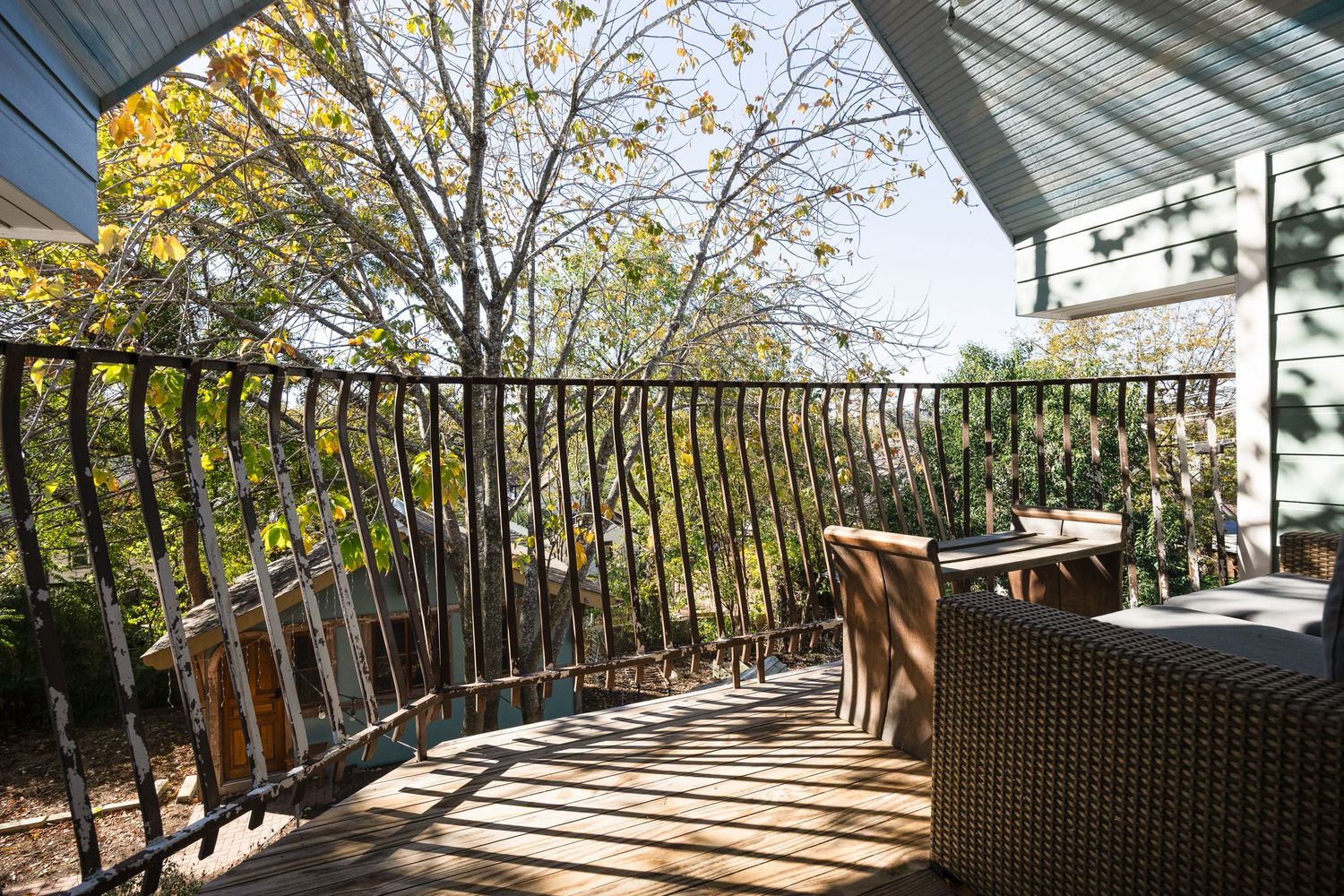 Sunny outdoor deck with wooden furniture and iron railings overlooking autumn trees.