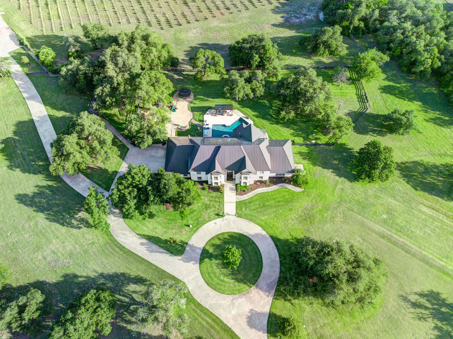 Aerial view of a sprawling estate with circular driveway, large home, pool, and lush green grounds surrounded by trees.