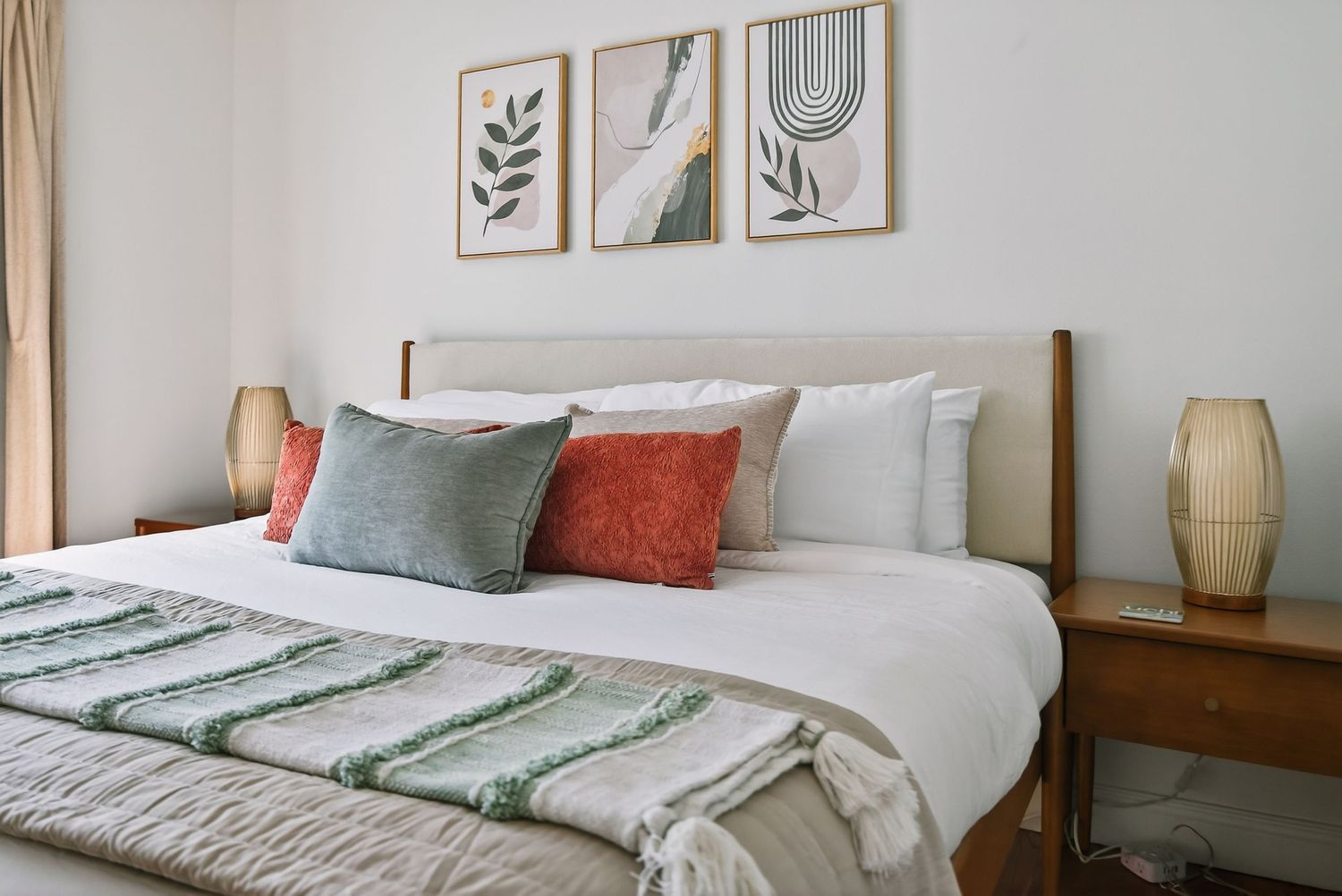 Modern bedroom with white bedding, colorful throw pillows, wooden furniture, and botanical artwork on the wall.