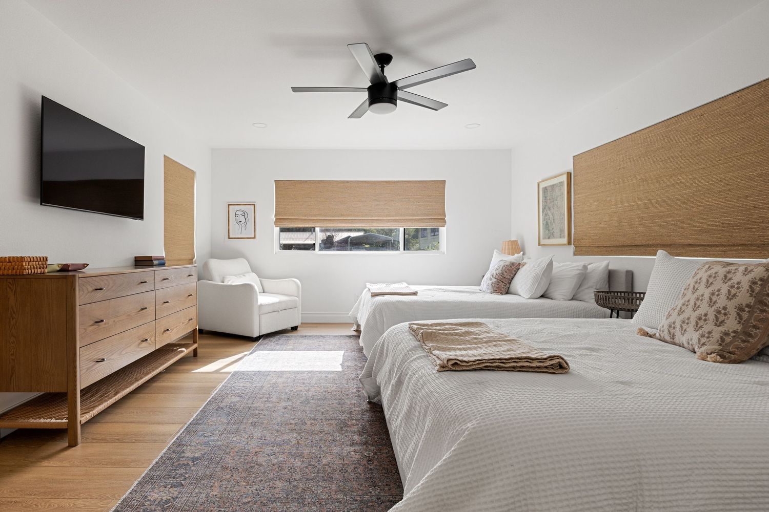 Bright master bedroom with two queen beds, wood headboard, ceiling fan, wall-mounted TV, and hardwood floors.