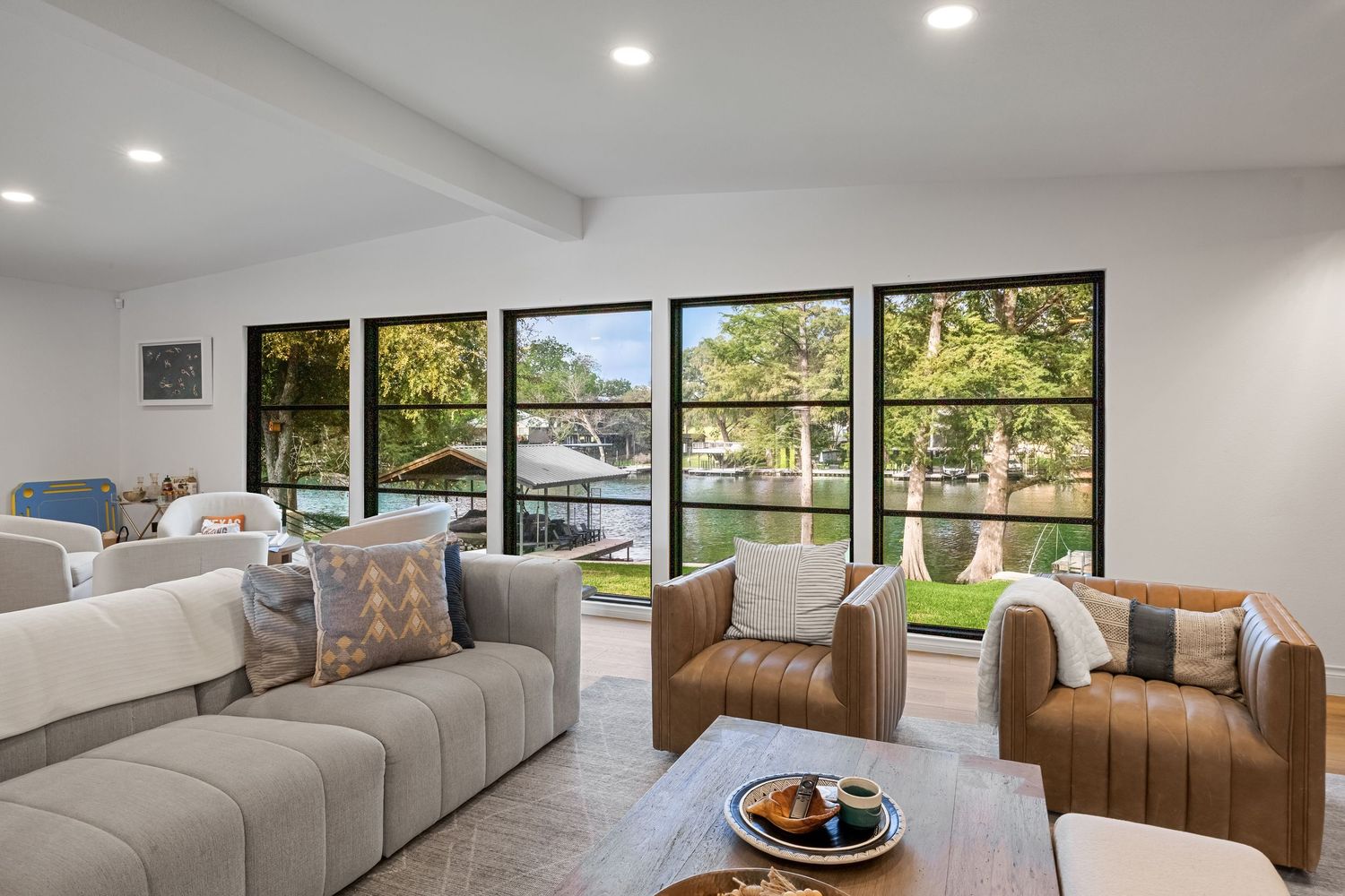 Modern living room with waterfront views, featuring leather chairs, gray sofa, and floor-to-ceiling windows overlooking a lake.