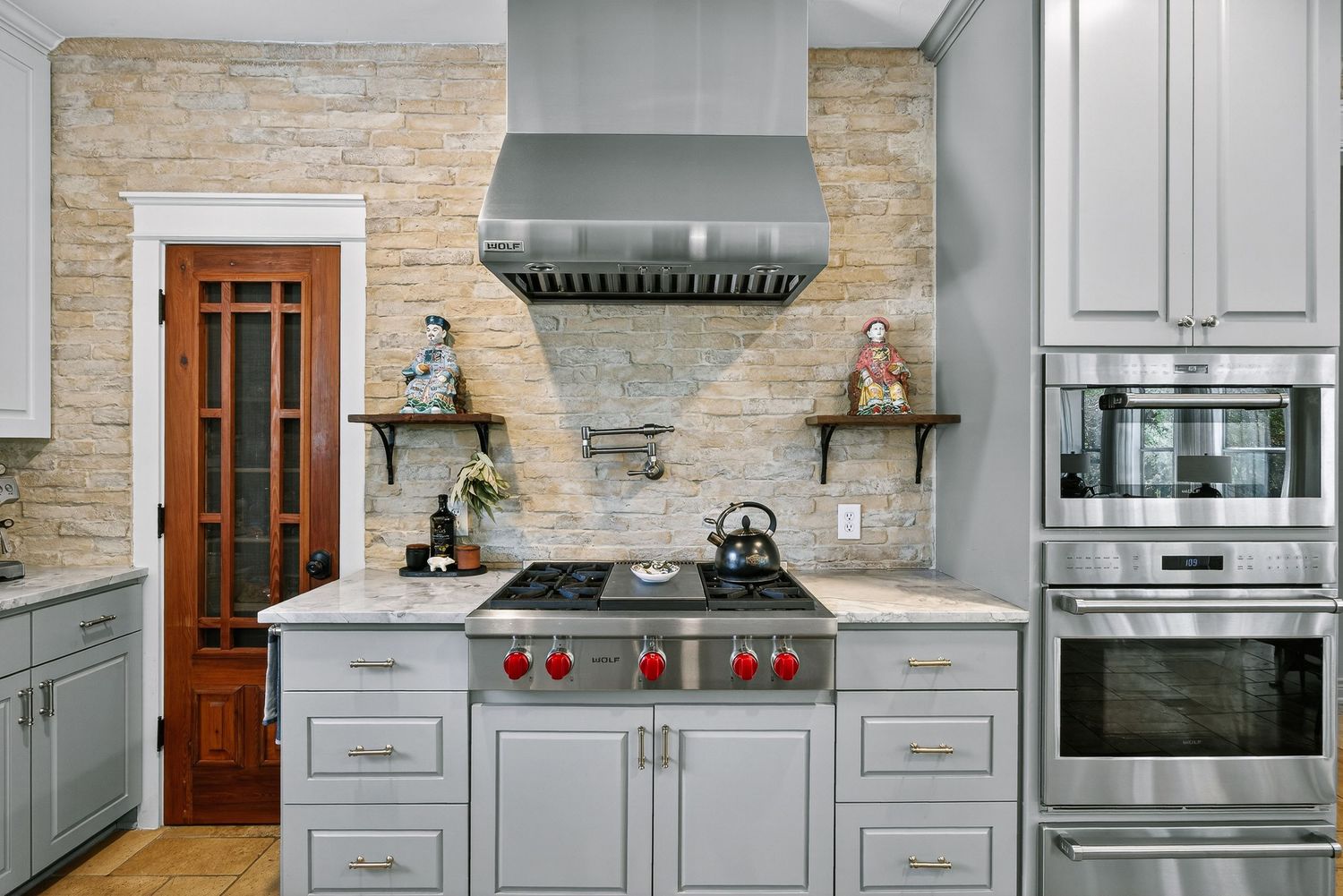 Modern kitchen with gray cabinets, stone backsplash, stainless steel range hood, and professional gas stove with red knobs.